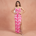 A woman models a white sleeveless maxi dress with a vibrant pink floral print, featuring thin straps, a ruched sweetheart bodice, and a high side slit. She is posing against a textured peach-pink studio backdrop.