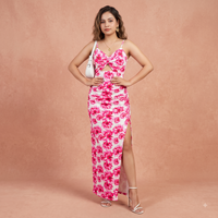 A woman models a white sleeveless maxi dress with a vibrant pink floral print, featuring thin straps, a ruched sweetheart bodice, and a high side slit. She is posing against a textured peach-pink studio backdrop.